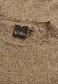 Brun stickad tröja med rund hals, med en subtil textur. Etiketten visar "Oscar Jacobson," med storlek M, tillverkad i Italien.