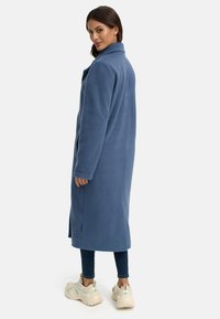 Blå fleecejacka med skräddarsydd design, sidoflikar och logotypdetalj på ärmen. Kombinerad med mörka jeans och kraftiga sneakers.