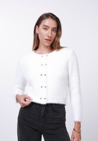 Λευκό κοντό πλεκτό ζακέτα από απαλό, αφράτο ύφασμα με στρογγυλή λαιμόκοψη και κουμπιά σε σχήμα καρδιάς. Συνδυασμένο με μαύρο ψηλόμεσο τζιν παντελόνι.