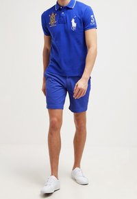 Blå polo med vit krage, broderad logotyp och nummerdetalj; tillsammans med matchande blå shorts och vita sneakers.