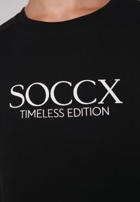 Camiseta negra de algodón con texto blanco en negrita que dice "SOCCX" y "EDICIÓN TIMLESS". Cuello redondo, textura suave.