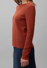 Pullover in Rostfarbe, strukturierter Strick, Rundhalsausschnitt, lange Ärmel, leicht lockere Passform, kombiniert mit navy-blauem Rock, kleiner Metallakzent am Saum.