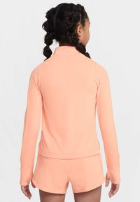 Dziewczyna z zaplecionymi włosami, ubrana w brzoskwiniowy top z długim rękawem i pasujące szorty, stoi tyłem na białym tle.