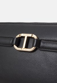 Dettaglio di una borsa in pelle nera con una chiusura in metallo dorato e incisione "TWINESET", superficie testurizzata, completata da una tracolla liscia.