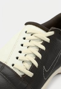 Καφέ δερμάτινο και εκρού πάνινο αθλητικό παπούτσι με λευκά κορδόνια, με λογότυπο στο πλάι και αντίθετη ραφή κατά μήκος του άνω μέρους.