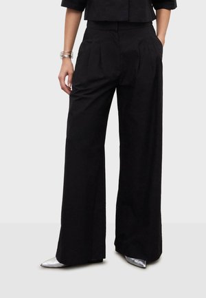 Personne portant un pantalon large noir, un haut court noir, des chaussures argentées pointues et un bracelet argenté, se tenant devant un fond uni.