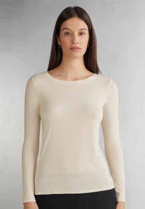 Beige Langarmshirt mit Rundhalsausschnitt, aus weichem Stoff, mit glatter Textur und taillierter Passform, getragen über schwarzen Hosen.