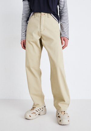 Beige cotton trousers with a straight leg, high waist, and front button. Paired with a dark blue striped long-sleeve shirt and light shoes.