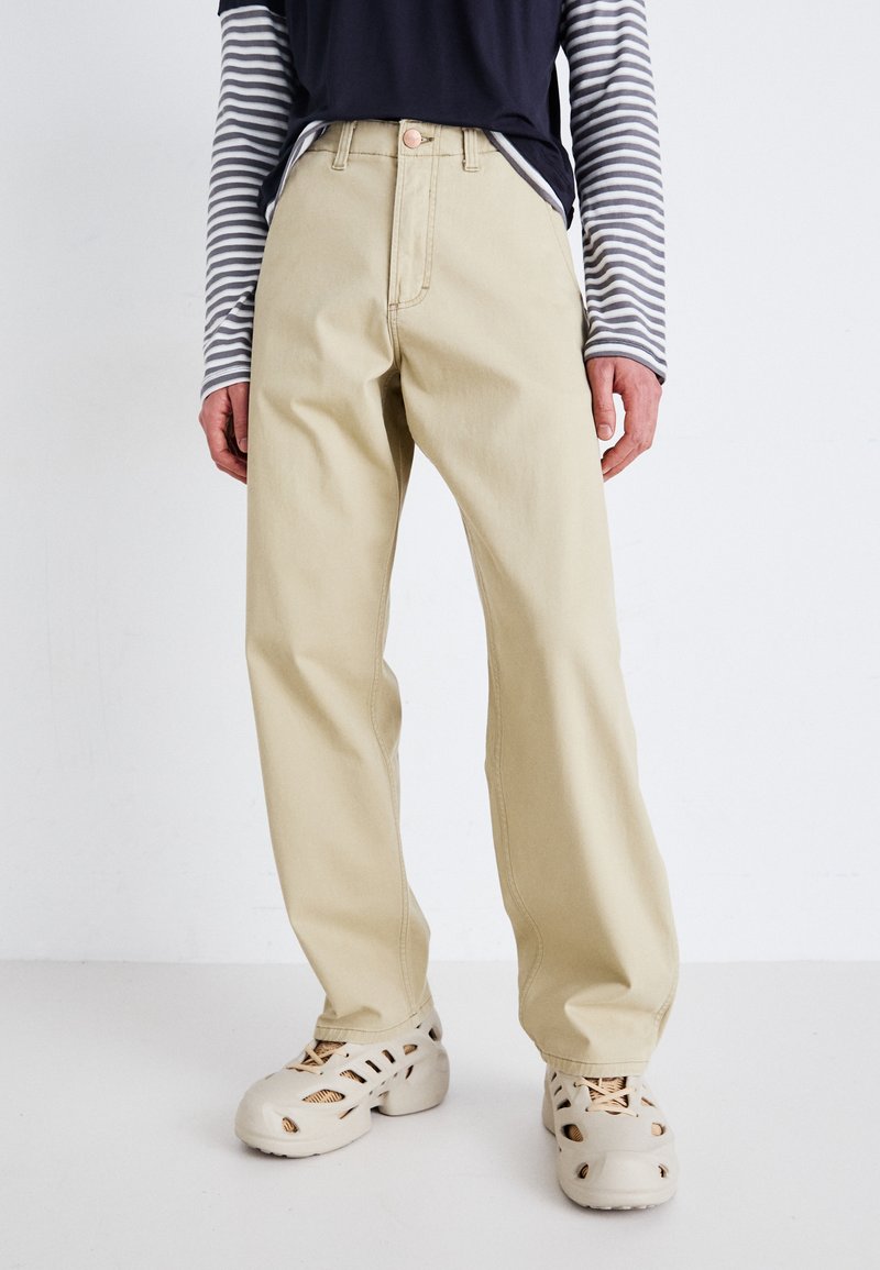 Pantalons en coton beige à jambes droites, taille haute, avec bouton à l'avant. Associés à une chemise à manches longues rayée bleu foncé et des chaussures claires.