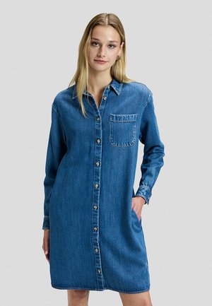 Jeune femme portant une robe chemise en jean bleu avec manches longues et une poche poitrine, debout avec une main dans la poche.