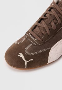 Brown suede sneaker with stitching details, a beige suede accent, and a white Puma logo. Features a textured toe and lacing system.