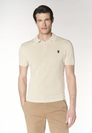 Uomo in piedi con le mani in tasca, indossa una polo beige a maniche corte con logo ricamato e pantaloni marrone chiaro.