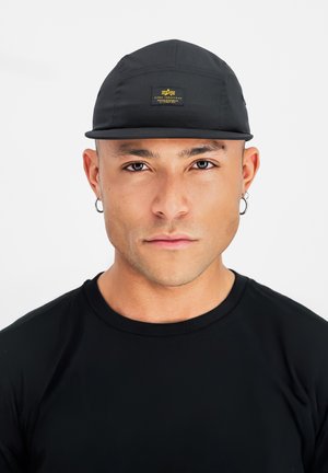 Young man with tan skin wearing a black cap, black shirt, and silver hoop earrings, looking straight ahead with a neutral expression.