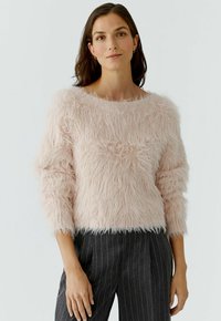 Vrouw met donker haar, gekleed in een lichtroze pluizig sweater en donkere gestreepte broek, staande tegen een effen witte achtergrond.