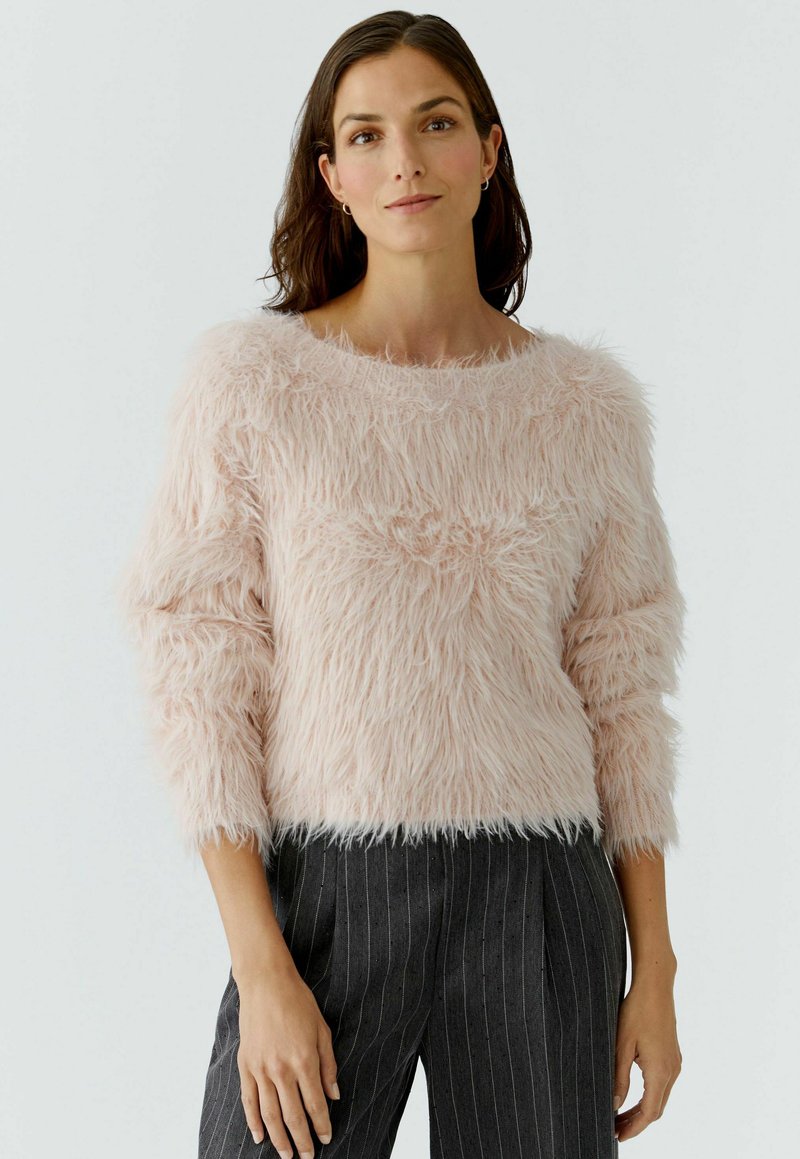 Vrouw met donker haar, gekleed in een lichtroze pluizig sweater en donkere gestreepte broek, staande tegen een effen witte achtergrond.
