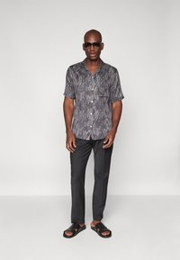 Chemise à manches courtes avec un motif imprimé serpent, gris et noir, assortie à un pantalon sombre et des sandales noires, portée par un homme.