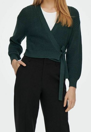 Femme portant un pull en maille vert foncé à nouer sur un haut blanc et un pantalon noir taille haute, une main dans la poche et l'autre détendue.