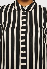 Camicia a righe bianche e nere con colletto, caratterizzata da righe larghe e strette alternate, e bottoni neri.