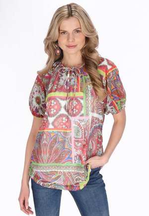 Kvinde med langt blondt hår iført en farverig blomstret bluse med puffede korte ærmer og blå jeans, poserer med den ene hånd i lommen.