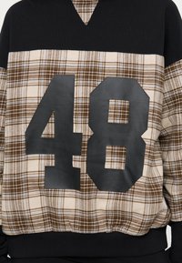 Ύφασμα με καρό μοτίβο σε μαύρο και καφέ χρώμα με μεγάλο μαύρο αριθμό "48" τυπωμένο στο κέντρο της μπροστινής πλευράς ενός πουλόβερ.