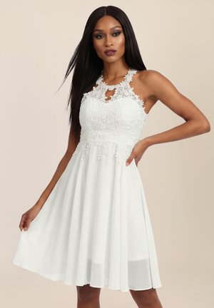 Frau trägt ein ärmelloses weißes Spitzenkleid mit Blumendetails, hält den Rock mit einer Hand und die andere Hand in der Hüfte.