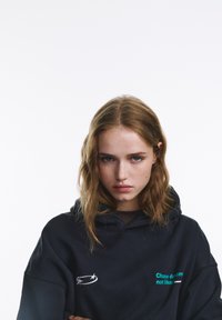 Fekete, laza szabású kapucnis pulóver fehér csillag grafikával és türkizkék felirattal: "Álmodj, ne úgy, ahogy mások." Lágy tapintású, hétköznapi dizájn.