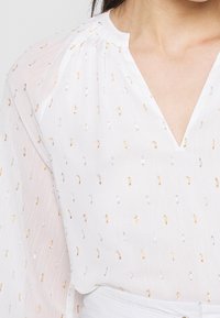 Blusa bianca con scollo a V e tessuto testurizzato. Presenta piccoli motivi metallici oro e argento su tutta la superficie. Maniche lunghe e trasparenti.
