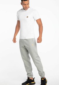 Camiseta blanca de manga corta con bolsillo y logo, combinada con pantalones de chándal gris claro, que tienen puños elásticos y zapatillas negras con acentos amarillos.