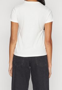 Bílá bavlněná tričko s krátkými rukávy, kulatým výstřihem a přiléhavým střihem, v kombinaci s černými kalhotami s širokými nohavicemi a zadními kapsami.