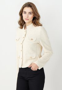 Beige Tweed-Jacke mit goldenen Knöpfen, zwei aufgesetzten Taschen und rundem Ausschnitt. Strukturierter Stoff und lange Ärmel.