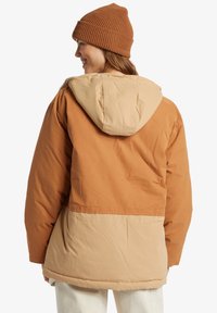 En puffjacka i brunt och beige med huva, med en slät yta och en kontrasterande fåll. En stickad brun mössa bärs på huvudet.