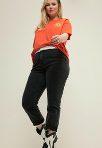 T-shirt arancione a maniche corte con grafica del sole, abbinato a jeans neri slim-fit con orli arrotolati e stivaletti alla caviglia in bianco e nero.
