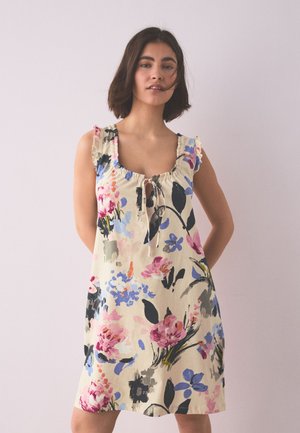 Frau mit kurzen dunklen Haaren, die ein ärmelloses beiges Kleid mit rosa, blauen und schwarzen Blumenmustern trägt, steht vor einer schlichten hellen Wand.