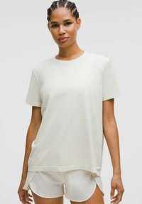 Vrouw met gevlochten haar in een losse witte korte mouwenshirt en textuur witte shorts tegen een effen grijze achtergrond.