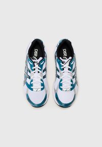 Baskets de sport blanches avec des accents turquoise, texture en maille, doublure intérieure noire et détails argentés réfléchissants. Caractéristiques des lacets blancs et un logo.