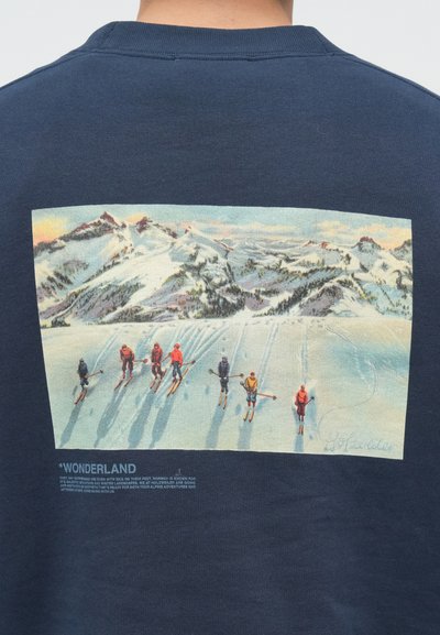Temno modra majica z okrasnim tiskom barvitega zimskega pokrajina na hrbtni strani, ki prikazuje smučarje ob snežnih gorah in opisno etiketo.