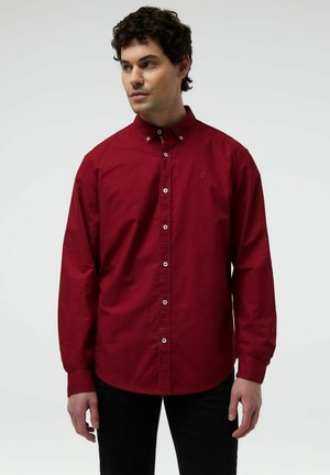 Mężczyzna w długim, czerwonym koszuli na guziki z kołnierzykiem zapinanym na guziki, stojący na jednolitym jasnym tle.