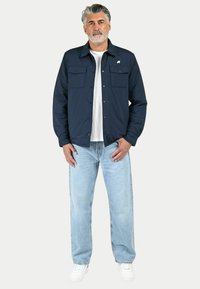 Giacca blu navy con due tasche sul petto, chiusura a bottoni, indossata sopra una maglietta bianca. I jeans azzurri chiari e le scarpe bianche completano il look.