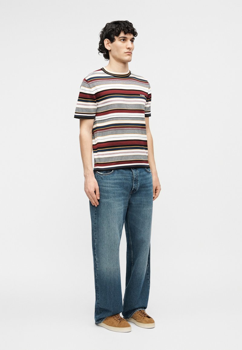 Young man standing wearing a multicolored striped short-sleeve shirt, loose blue jeans, and brown sneakers against a plain background.