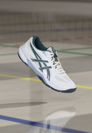 Scarpa da ginnastica bianca e grigia con lacci, che fluttua sopra un pavimento di palestra segnato con linee colorate.