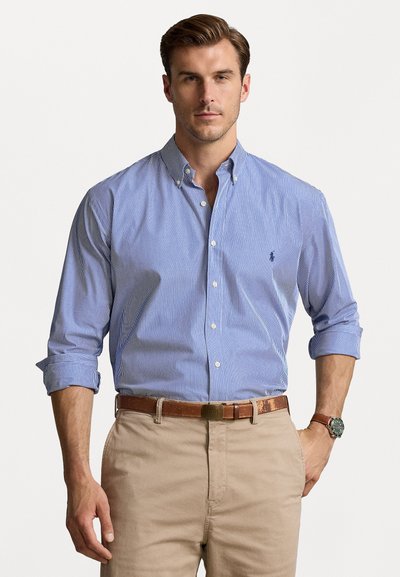 Una camicia a righe blu e bianche con colletto e maniche lunghe è abbinata a pantaloni beige e una cintura marrone. È indossato un orologio da polso.
