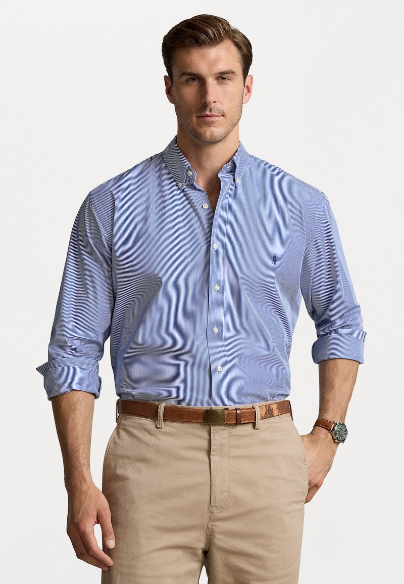 Une chemise à rayures bleues et blanches, à col et à manches longues, associée à un pantalon beige et une ceinture marron. Une montre-bracelet est portée.