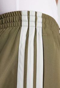 Pantaloni corti verde oliva con una vita texturizzata, tre strisce verticali bianche e un tessuto liscio con sottili dettagli di cucitura.