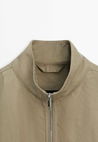 Colletto di giacca beige con cerniera argentata visibile, colletto corto a listino piegato e cuciture ordinate su sfondo bianco uniforme.