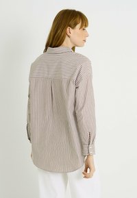 Camisa de botones a rayas en beige y marrón. Presenta un corte relajado, mangas largas y un pliegue en la espalda para mayor movilidad.