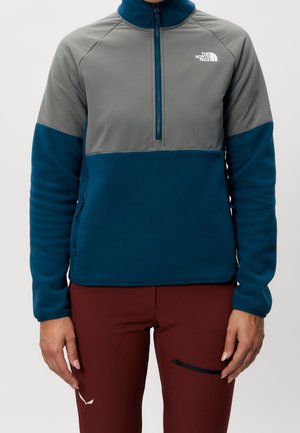 Person trägt einen blau-grauen Viertelzip-Fleece mit The North Face-Logo, kombiniert mit bordeauxroten Hosen mit Reißverschlusstasche am Oberschenkel.