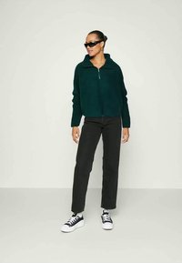 Jachetă din fleece, cu fermoar, de culoare verde închis, având un guler înalt. Asortată cu blugi negri cu croială dreaptă și adidași negri cu alb.