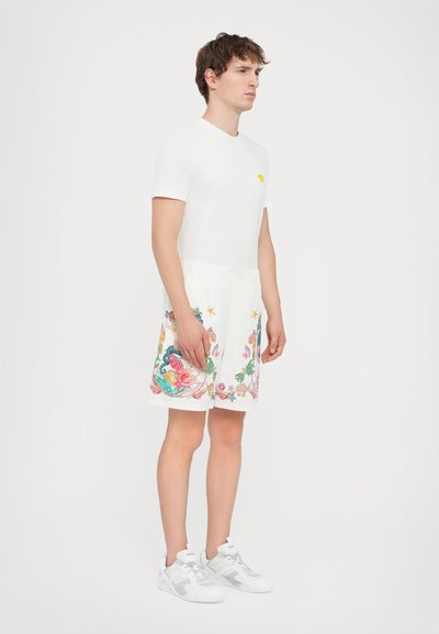 White cotton t-shirt with a small yellow logo, paired with elastic waist shorts featuring colorful seashell and marine designs. White sneakers.