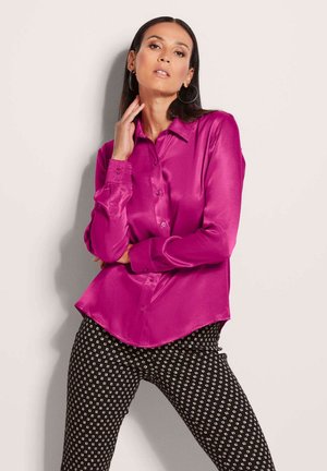 Fuchsia Satinbluse mit knöpfbarem Kragen, tailliertem Schnitt und langen Ärmeln, kombiniert mit gemusterten schwarzen Hosen mit weißen Quadraten.