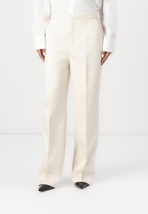 Pantaloni su misura color crema con design a gamba dritta, caratterizzati da tasche laterali e una superficie liscia e texture. Abbinati a scarpe nere a punta.